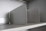 Gray metal shelf dividers are attached to a shelf, forming two vertical partitions. The dividers have rounded corners and small holes near the edges, and they are positioned against a plain background.