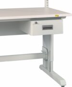 A gray metal workbench with an attached drawer beneath the tabletop. The bench has an adjustable support frame, visible legs, and a small yellow warning label on the corner of the tabletop.