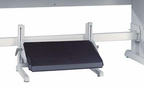 A metal footrest with a black, textured, rectangular surface adjustable by a lever is placed under a desk. The footrest is designed to support foot placement and improve ergonomic posture while seated.