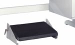 A black and silver ergonomic footrest with an inclined surface is positioned on a light-colored floor. The footrest features a ridged texture on its top surface and has metal supports on the sides.