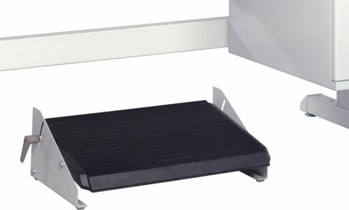 A black and silver ergonomic footrest with an inclined surface is positioned on a light-colored floor. The footrest features a ridged texture on its top surface and has metal supports on the sides.