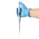 A gloved hand holds a white pipette against a white background. The pipette has measurement markings, a digital display, and a tip ready for liquid transfer, typical in laboratory settings.