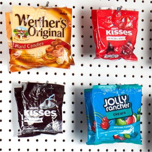 Four candy packages hanging on a pegboard: Werthers Original Hard Candies (orange), Hersheys Kisses Milk Chocolate in both red and black, and Jolly Rancher Chews (blue), all secured with the Vulcan SP HOOK 4 MEDIUM W/BALL from the 10PK.