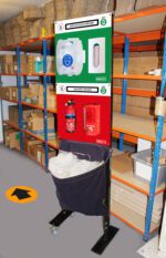 A safety station in a warehouse with shelves behind it. The station includes a mask and glove dispenser on top and a fire safety section with an extinguisher and alarm below. A bin with transparent bags is underneath. A yellow arrow is on the floor.