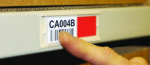 A finger pointing at a white label with a barcode and the text CA004B on a shelf. A red square is next to the label. The background includes a blurred cardboard box.
