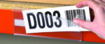 A person inserts a paper label with the code D003 and a barcode into a transparent holder on an orange shelf edge, enhancing warehouse designs by streamlining inventory management.