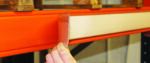 A hand is expertly placing a transparent label holder on an orange metal beam, part of innovative shelving or racking in a warehouse. This detail-oriented action reflects efforts to improve warehouse designs. A wooden pallet rests securely on top of the beam.