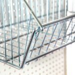 A Vulcan WIRE 12 OEB DIVIDE shopping basket hangs on a pegboard wall. The shiny silver wires form a neat grid pattern with visible angled edges, and the boards spaced holes are ideal for organization. (10 per carton).