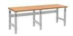 A long, rectangular workbench with a light brown wooden top and adjustable metallic legs. The legs are gray and have multiple holes for height adjustment. The bench is set against a plain white background.
