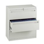 A white metal filing cabinet with two drawers. The top drawer is closed and locked, while the bottom drawer is partially open, revealing organized files. The cabinet is set against a white background.