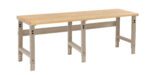 A long, sturdy workbench with a light wooden top and metal legs. The frame is painted in a beige color. The bench is designed for industrial or workshop use and rests on a solid surface.