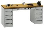 A metal workbench with a wooden top is shown, featuring several drawers on both sides. Various tools, including a drill, a wrench, and a clamp, are placed on the bench. A vise is mounted on the right side of the work surface.