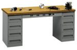 A sturdy workbench with a wooden top and two sets of metal drawers on each side. Tools are scattered on the surface, including a drill, a clamp, and wrenches. The drawers have handles for easy access.