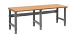 A long wooden workbench with a light brown top and dark gray metal adjustable legs. The design is simple and functional, suitable for workshops or garages. The bench has sturdy metal crossbars for added support.