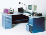 A retro L-shaped desk with blue drawers, featuring an old computer monitor, keyboard, black desk lamp, files, a camera, framed picture, a paper tray, and a glass. The desk is set against a plain white background.