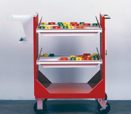 A red utility cart with two shelves, each holding colorful, rectangular objects. The cart has wheels for mobility and a white attachment on one side. The background is a plain white wall with a gray floor.