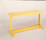 A bright yellow metal table with a flat rectangular top and sturdy angled legs, set against a plain white background. The table has a simple, industrial design.