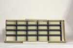 An empty, beige metal shelving unit with multiple compartments and black dividers sits against a plain white background. A single white tube lies in front of the shelving.