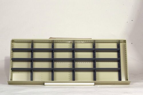 An empty, beige metal shelving unit with multiple compartments and black dividers sits against a plain white background. A single white tube lies in front of the shelving.