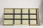 A beige metal shelving unit with a grid-like structure and several black horizontal dividers, standing against a plain white background. A fluorescent light tube sits at the base of the unit.