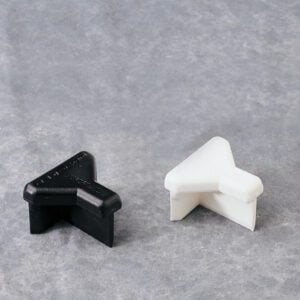 A black and a white plastic corner protector, shaped like a right angle, are placed on a light gray textured surface. They are designed to protect furniture or walls from damage.
