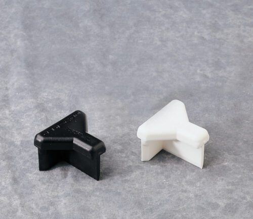 A black and a white plastic corner protector, shaped like a right angle, are placed on a light gray textured surface. They are designed to protect furniture or walls from damage.