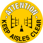 A circular yellow sign with black text reads Attention Keep Aisles Clear, featuring a graphic of shelves with an empty aisle, similar to MH-USA Floor Graphic Markers - 16.93 in. dia.