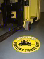 On the gray floor, a yellow sign labeled Attention: MH-USA Floor Graphic Markers - 16.93 in. dia. - Forklift Truck Area with a forklift graphic warns of the nearby yellow forklift, highlighting the caution required in this zone.