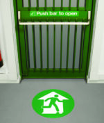 A green emergency exit door with a push bar features an MH-USA 16.93 in. dia. Floor Graphic Marker, displaying a white figure running towards an open door, indicating the fire exit direction.