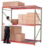 A person arranges large brown cardboard boxes on a red and gray metal shelving unit beside wire mesh cages. The shelves are partially filled, and one large box is placed on the floor. The background is plain white.