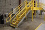 A yellow metal staircase with side railings leads to an elevated platform, forming part of a custom catwalk system in an industrial setting. The area features concrete walls and floor, with a fire extinguisher prominently displayed on the wall.