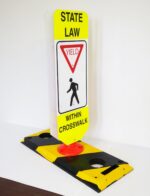 The TCT Flexible Post Crosswalk System, State Law - Yield features a sign on a yellow/black base that reads State Law: Yield to Pedestrians Within Crosswalk, with a pedestrian symbol and yield triangle, set against a plain white background for enhanced visibility.