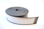 A roll of MH-USA Magnetic Label Holder tape, flat and flexible, is slightly uncoiled on a white surface. The dark gray material contrasts with its distinctive white stripe on one side and measures H.2.36 in. x W3.94 in.