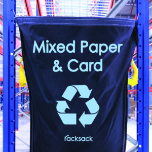 A blue MH-USA Racksack - Mixed Paper & Card hangs on metal shelving in a warehouse, featuring a white recycling symbol and the Racksack brand name at the bottom.