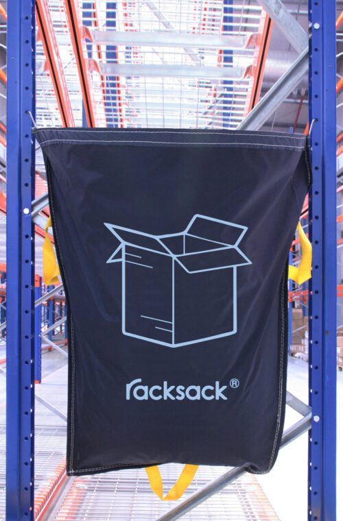 RSB1CNT.jpg A large black MH-USA Racksack Symbol Only - Cardboard with a blue outline and bold racksack print is attached to blue metal shelves in a warehouse, hanging amidst orange and blue shelving units. In the background, cardboard boxes are neatly stacked.