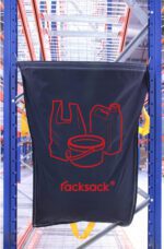 A large black MH-USA Racksack Symbol Only - Plastic Only, adorned with red waste symbols such as a plastic bag, container, and can, hangs on a blue industrial rack in a storage facility. Its yellow handles enhance the designs efficiency.