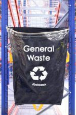 A clear MH-USA Racksack labeled General Waste with a recycling symbol hangs on a blue industrial racking system, set against metal shelves in a busy warehouse, showcasing the quality of MH-USA Racksack solutions for waste management.