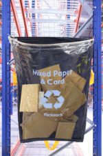 An MH-USA Racksack Clear - Mixed Paper & Card is suspended on a blue rack, filled with cardboard pieces. The setting shows a warehouse with shelves and a prominent red metal framework.