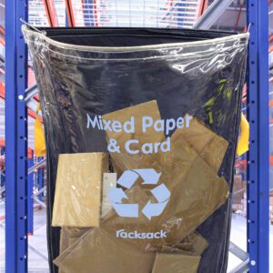 An MH-USA Racksack Clear - Mixed Paper & Card is suspended on a blue rack, filled with cardboard pieces. The setting shows a warehouse with shelves and a prominent red metal framework.