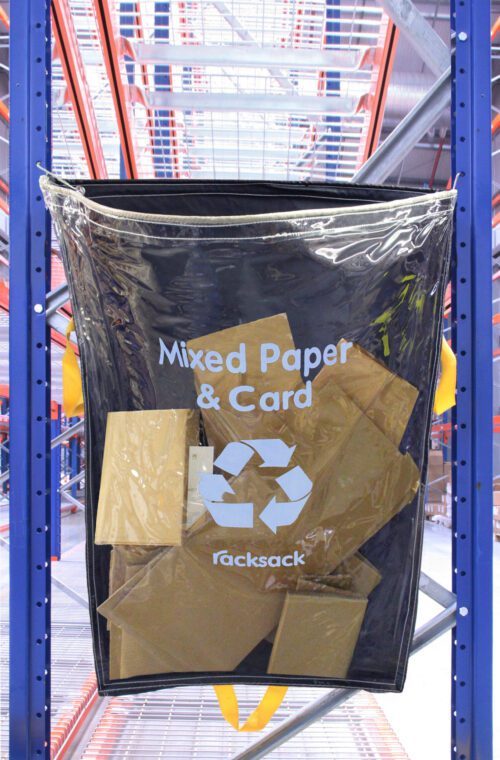 An MH-USA Racksack Clear - Mixed Paper & Card is suspended on a blue rack, filled with cardboard pieces. The setting shows a warehouse with shelves and a prominent red metal framework.