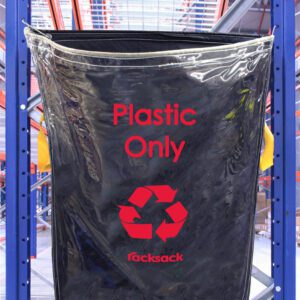 A blue and black MH-USA Racksack Clear - Plastic Only bag hangs on a metal rack in a warehouse. It displays a red recycling symbol and racksack at the bottom, with orange-beamed shelving and metal grating visible in the background.