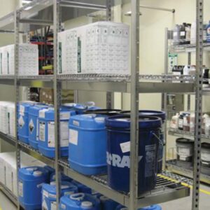 Metal shelves in a storage room hold various containers, including large blue and white canisters and boxes. The shelves are organized with clear labeling, and the background displays additional shelving with more containers.