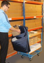 In a storage area, someone in glasses and a blue jacket arranges flat boxes into a covered MH-USA Racksack Trolley - Double Pocket. Behind them are shelves of neatly stacked cardboard boxes.