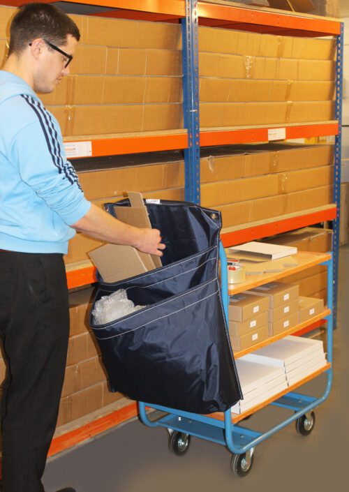 In a storage area, someone in glasses and a blue jacket arranges flat boxes into a covered MH-USA Racksack Trolley - Double Pocket. Behind them are shelves of neatly stacked cardboard boxes.
