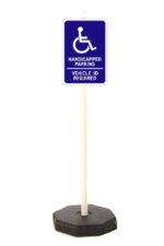 A blue handicapped parking sign with a wheelchair symbol and HANDICAPPED PARKING VEHICLE ID REQUIRED text on a white pole secured in a durable TCT Octagon Sign Base, 30Lbs.