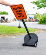 A hand holds a vibrant orange Caution Work Zone Ahead sign on a TCT Heavy Duty 60lb base with 1/2 steel axle and rubber wheels, sturdily placed on a paved path. Industrial equipment and lush greenery form the backdrop.