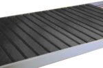 A close-up of an empty black conveyor belt with metal edges. The surface is segmented with parallel grooves, and it appears to be made of rubber or a similar material. The background is white and out of focus.