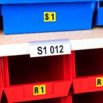 Blue storage bins labeled S1 and red ones with R1 stickers are neatly arranged on shelves. An MH-USA Angled self-adhesive Ticket Holder, 1.02 in. x 3.94 in., Pk 100 decorates a shelf bearing the label S1.
