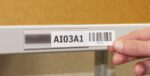 A hand applies a label marked AI03A1 and a barcode into an MH-USA Angled self-adhesive Ticket Holder 1.54 in. x 3.94 in. Pk 100 on a shelf, with a cardboard box visible in the background.