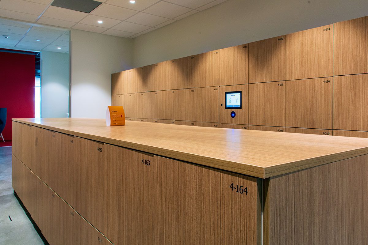 A room with wooden lockers, each numbered, fills the space. A small control panel is embedded in these smart locker systems, and a bright orange item rests on the surface in the foreground. The walls and ceiling are light-colored.